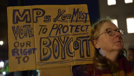 A-protestor-holds-a-cardboard-placard-that-reads,-“MPs-you-must-vote-to-protect-our-right-to-boycott”-during-a-nighttime-pro-Palestine-protest-outside-the-Houses-of-Parliament