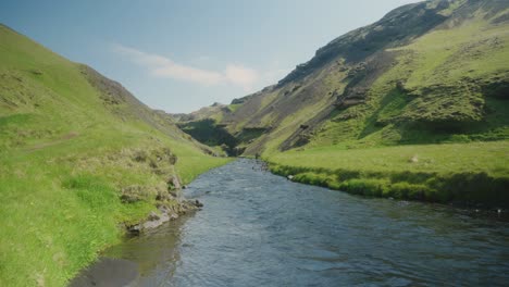 Ein-Kleiner-Fluss-Und-Moosige-Hügel-In-Island,-Sonnige-Grüne-Felsen-Und-Blaues-Wasser-Im-Vordergrund
