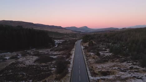 Twilight-over-a-solitary-road-through-rugged-terrain,-snow-patches,-serene-landscape,-aerial-view