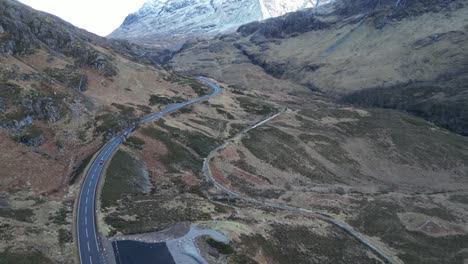 Winding-road-in-Glencoe-with-snow-capped-mountains-in-the-distance,-overcast-weather,-aerial-view