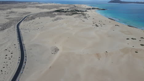 Rising-aerial-view-over-Corralejo-Beach-Fuerteventura-empty-curving-road-winding-across-sandy-beach-towards-the-coastline