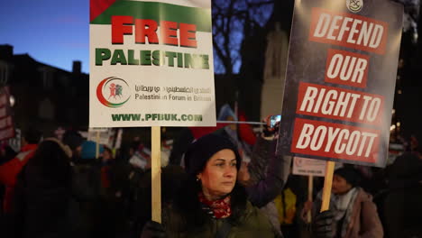 A-protestor-holds-up-two-placards-that-read,-“Free-Palestine”-and,-“Defend-our-right-to-boycott”-during-a-nighttime-protest-outside-the-Houses-of-Parliament