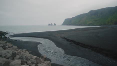 River-flows-to-meet-the-ocean-at-Víkurfjara-Black-Sand-Beach-in-Vik,-Iceland,-Reynisfjall-in-the-distance