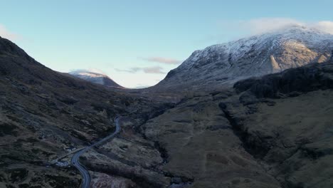 Glencoe-Valley-with-a-winding-road-amidst-rugged-snow-capped-mountains-at-dusk,-aerial-view