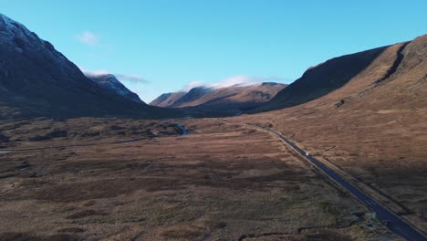 Glencoe-valley-with-a-winding-road-amidst-scenic-mountains,-clear-blue-sky,-aerial-view
