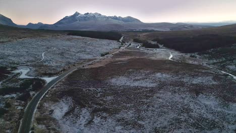 Snowy-landscape-in-Skye-with-a-winding-road-and-mountain-backdrop,-twilight,-aerial-view