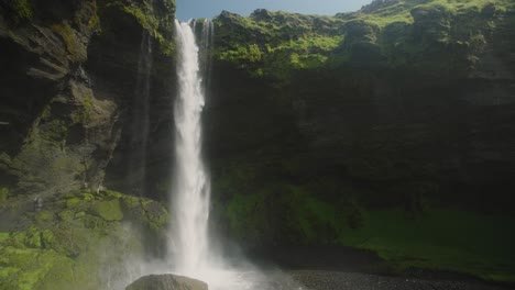 Kvernufoss-waterfall-in-Iceland,-sunny-green-rocks-in-the-foreground-and-small-figures-hiking-underneath
