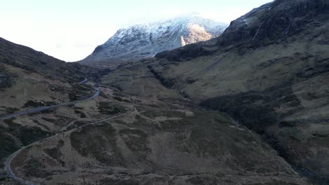 Winding-road-through-Glencoe-with-snow-capped-mountains,-aerial-view-at-dusk