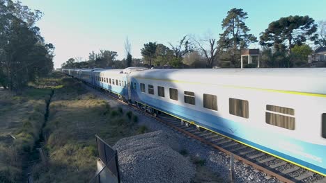 Passenger-trains-with-many-cars-drive-along-tracks-behind-homes-in-countryside
