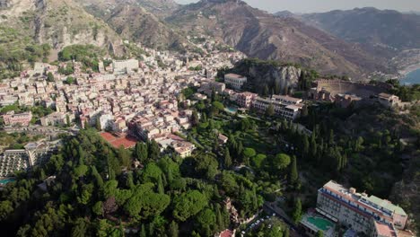 Aerial-drone-video-over-Taormina,-Sicily-capturing-the-old-town-buildings-and-green-trees-and-mountains-in-the-background