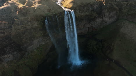 Aerial-view-backwards-away-from-a-waterfall,-sunny,-fall-day-in-Iceland