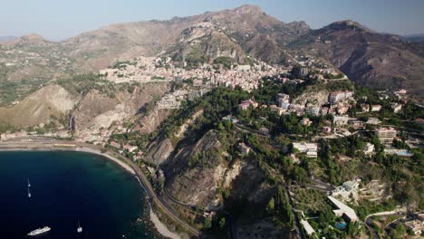 Drohne-Fliegt-über-Den-Ozean-In-Richtung-Einer-Stadt,-Die-Auf-Dem-Gipfel-Des-Berges-Namens-Taormina-Erbaut-Wurde