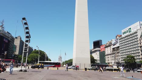 People-walk-around-Obelisk-Landmark-in-9-de-Julio,-Corrientes-Avenue-City-Center-Tourists-enjoy-Downtown-Travel-Street-Location,-Traffic-driving-by