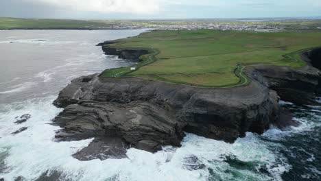 Waves-breaking-along-Kilkee-cliffs-with-coastal-village-in-background,-County-Clare-in-Ireland