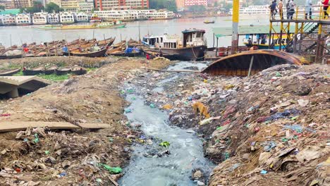 Sewage-pollution-in-the-river,-garbage-on-the-riverside-with-fishing-boats-and-modern-buildings-in-the-background,-Dhaka,-Bangladesh