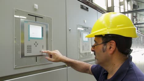 Middle-aged-man-with-white-beard-in-hard-hat-presses-control-panel-buttons-at-factory