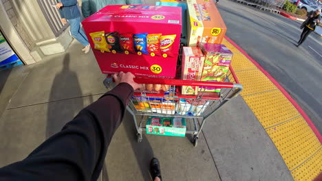 Close-up-shot-of-shopping-cart,-full-of-snacks-and-other-grocery-items-while-walking-outside-to-the-car-park-on-a-sunny-day