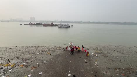 Rear-view-wide-shot-of-people-bathing-and-offering-prayers-to-god-near-Hooghly-river-with-cityscape-at-background-in-Kolkata,-India