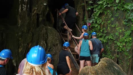 Tourists-wear-hard-hats-climbing-up-old-metal-stairs-entering-cave-tour-in-tropical-jungle