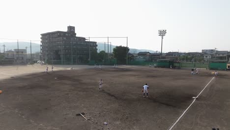 School-Baseball-Team-Trains-Playing-Match-at-Ground-Field,-Aerial-Slow-Motion-view,-Skyline,-Mountains-and-City-Background