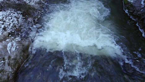 Aerial-close-up-crossing-along-the-mountain-river-with-a-small-fall-of-water-during-the-winter-with-some-ice-and-snow,-turbulent-water-,-rocky-shore