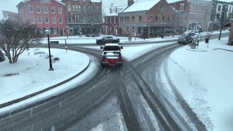 Pickup-truck-with-snowplow-and-salt-spreading-driving-around-small-town-square-covered-in-snow-and-Christmas-decorations