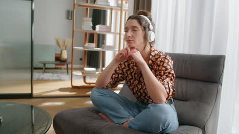 Woman-sitting-cross-legged-on-chair-wearing-headphones-listens-to-podcast