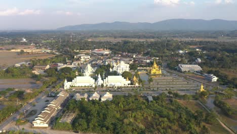 Luftdrohne-Von-Wat-Rong-Khun,-Dem-Riesigen-Buddhistischen-Weißen-Tempel-Und-Dem-Goldenen-Tempel-Mit-Bergen-Und-Landschaft-In-Chiang-Rai,-Thailand