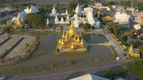 Aerial-drone-backwards-of-Wat-Rong-Khun-the-giant-buddhist-White-Temple-and-Golden-Temple-with-mountains-and-landscape-in-Chiang-Rai,-Thailand