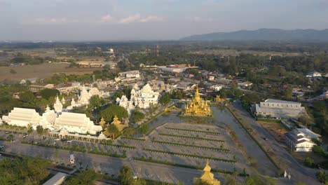Aerial-drone-circling-around-Wat-Rong-Khun-the-huge-buddhist-White-Temple-and-Golden-Temple-with-mountains-and-landscape-in-Chiang-Rai,-Thailand