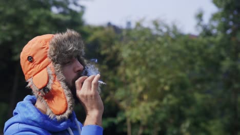 Man-in-a-blue-sweatshirt-is-smoking,-he-is-wearing-a-warm-orange-winter-aviator-hat