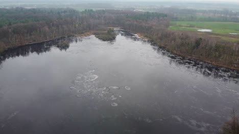 Aerial-view-over-lake-covered-with-ice-in-snowy-winter-forest-landscape-in-Retie,-Belgium