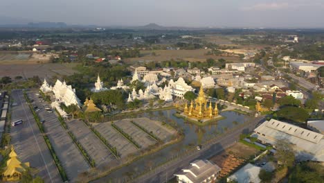 Aerial-drone-wide-view-of-Wat-Rong-Khun-the-huge-buddhist-White-Temple-and-Golden-Temple-with-mountains-and-landscape-in-Chiang-Rai,-Thailand