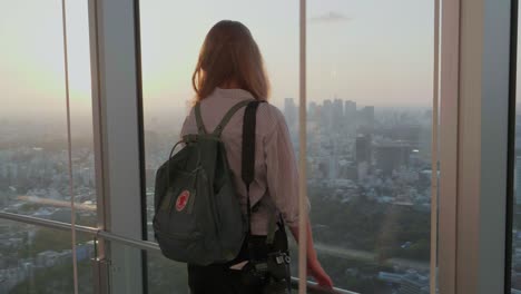 Lonely-blond-female-tourist-with-a-backpack-and-a-camera-is-standing-on-a-tower-overlooking-the-skyline-of-Tokyo,-Japan