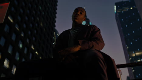 Afro-American-model-sitting-in-front-of-camera-in-smart-city-with-modern-skyscraper-building-illuminated-at-night-in-background