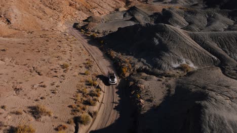 Aerial-tracking-shot-of-white-SUV-on-winding-desert-road-through-brown-hills