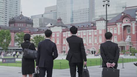 four-young-japanese-business-people-in-suits-walking-in-front-of-Tokyo-Station-in-Chiyoda,-Tokyo,-Japan-on-an-overcast-day