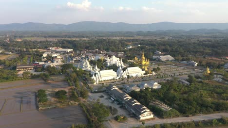 Aerial-drone-backwards-of-Wat-Rong-Khun-the-huge-buddhist-White-Temple-and-Golden-Temple-with-mountains-and-landscape-in-Chiang-Rai,-Thailand