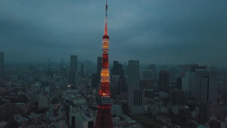 Drone-aerial-above-Tokyo-City-panning-towards-the-Tokyo-Tower-surrounded-by-tall-skyscrapers-at-dusk-with-moody-dark-overcast-clouds-and-smog-polluted-skies