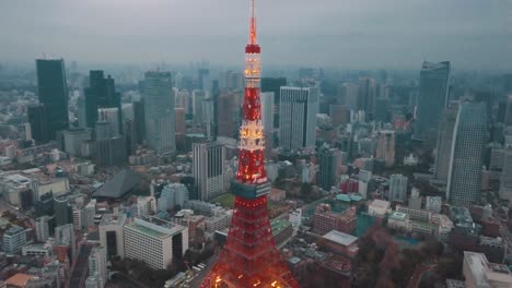 Drohnenantenne-über-Der-Stadt-Tokio,-Die-In-Der-Abenddämmerung-In-Richtung-Des-Von-Hohen-Wolkenkratzern-Umgebenen-Tokio-Towers-Schwenkt,-Mit-Stimmungsvollen-Dunklen-Wolken-Und-Einem-Smogverschmutzten-Himmel