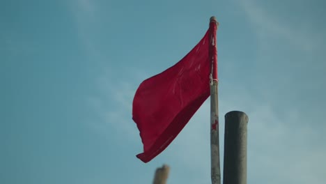 Tattered-red-flag-fluttering-against-a-clear-blue-sky,-symbolizing-distress-or-warning,-shot-in-daylight