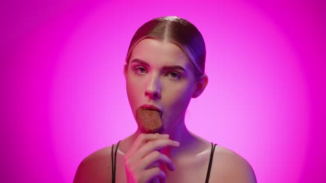Young-adult-blonde-caucasian-girl-eats-an-ice-cream-stick-in-a-pink-studio-with-chroma-background