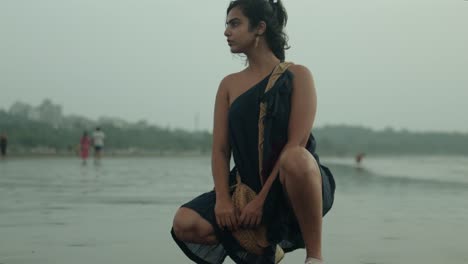 Woman-in-a-sari-squatting-on-a-wet-beach,-looking-off-to-the-side-with-wind-in-her-hair,-overcast-weather