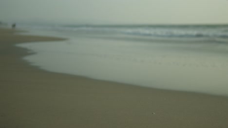 Neblige-Strandszene-Mit-Sanften-Wellen-Und-Einer-Einsamen-Figur-In-Der-Ferne,-Die-Ein-Gefühl-Der-Ruhe-Hervorruft,-Aufgenommen-Mit-Weichem-Fokus