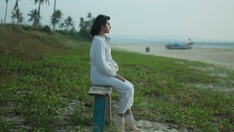 Woman-in-white-sitting-on-a-bench-by-the-beach-at-daylight,-looking-thoughtful,-with-a-boat-and-sea-in-the-background,-serene