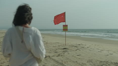 Red-warning-flag-on-an-empty-beach-with-young-women-walking,-overcast-weather