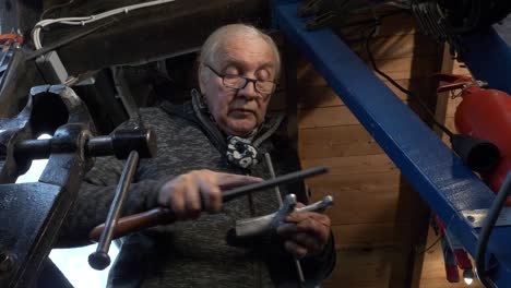 Senior-male-worker-uses-a-steel-file-on-a-machine-part,-shot-looking-up,-workshop-with-a-bench-vice