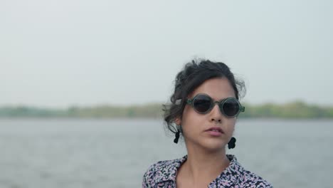Young-woman-with-sunglasses-by-the-water,-looking-away-thoughtfully,-with-a-soft-focus-on-the-background,-tranquil-vibe