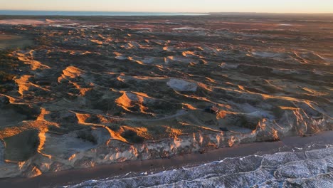 Aerial-view-of-large-sand-dunes-standing-by-the-sea-illuminated-by-the-sunset-light,-a-summer-house-stands-on-the-dunes,-and-sea-water-washes-the-coast