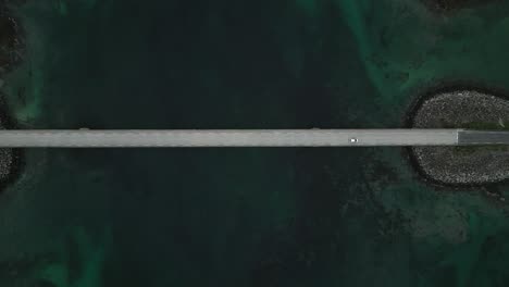 An-aerial-top-down-view-shows-a-car-driving-across-a-bridge-in-Lofoten,-Norway,-as-the-brilliant-turquoise-blue-hues-of-the-water-beneath-the-structure-captivate-the-scene